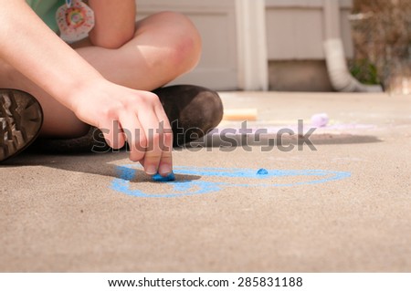 View of a child\'s hand holding blue sidewalk chalk and drawing on a driveway.  The child is sitting cross-legged and is drawing a heart with blue chalk.