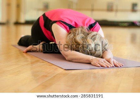 An older woman doing yoga practices pigeon pose. The blond woman is wearing a bright pink shirt and she is practicing on a mauve colored yoga mat. She is alone in a studio classroom.