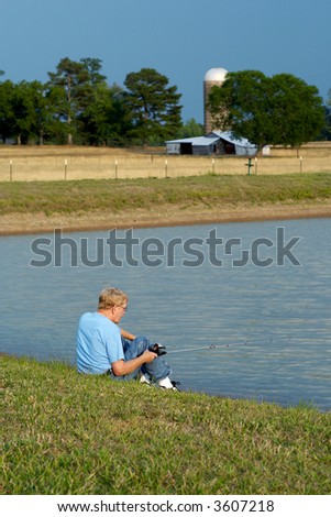 An older man sitting by the pond fishing with a pasture and old barn in the background.