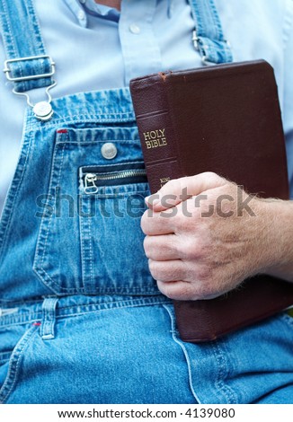 A man in overalls holding the Bible against his chest. Narrow depth of field. Focus is on his hand and Bible.