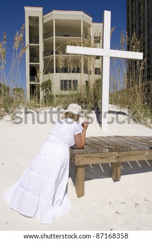 A woman kneeling and praying by a cross at the beach.