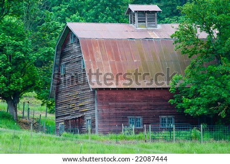 Old red wooden barn among green trees and field, roof, cupola, weathered, rustic.