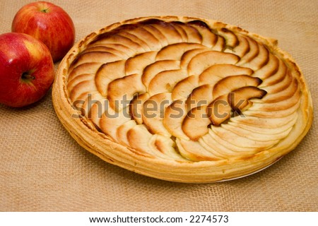 Fresh apple tart with two apples lying alongside over coarse jute fabric for a country look.