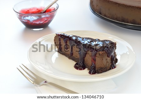 Slice of chocolate cheesecake with blackcurrant jam on white plate. Cheesecake made of cheese, cream, dark chocolate and some espresso and amaretto. Crust made of almond, biscuits and chocolate.