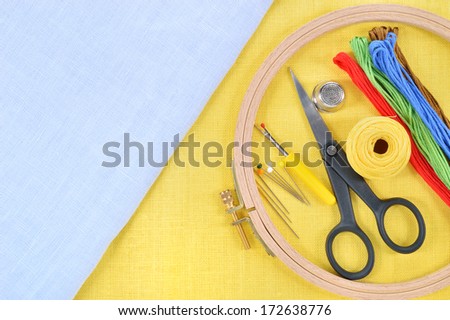Embroidery and cross stitch accessories on yellow and light blue linen fabric. Embroidery hoop, scissors, thread, needles, thimble. Copy space.