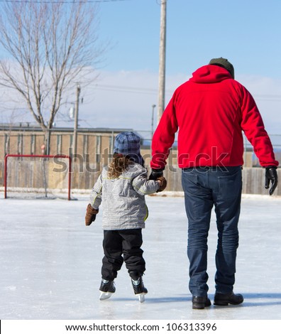Father teaching daughter how to ice skate at an outdoor skating rink in winter.