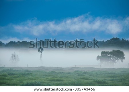 Farm windmill projecting above bank of fog on Australian farm under dark storm clouds and low white cloud