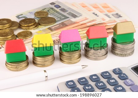 A rising stack of Euro coins with toy houses on top, cash, calculator and pen. A Euro house financial metaphor.