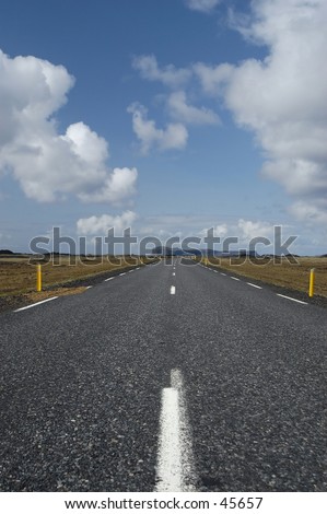 Road going into the distance, lots of blue sky and clouds above, mountains in the distance, yellow marker posts along the road.