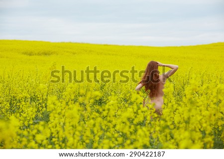 rapeseed field with yellow flowers, naked red-haired girl standing with her back