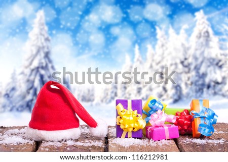 red Santa Claus hat and colorful christmas gifts on a wooden table with snow outside with an amazing winter wonderland in blurred background, forest in winter