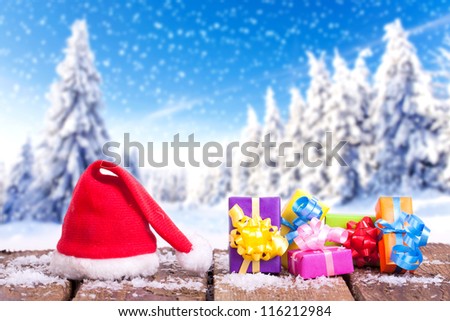 red Santa Claus hat and colorful christmas gifts on a wooden table with snow outside with an amazing winter wonderland in blurred background, forest in winter