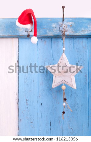 a red Santa Claus hat and various stars hanging on a rustic hook strip on blue wooden board, christmas decoration and christmas welcome
