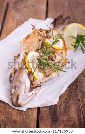 grilled trout with lemon pieces, fresh herbs and garlic on white baking paper on wooden table.