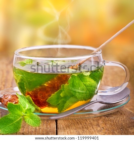 steaming hot tea in glass cup with mint leaf and brown cane sugar on a wooden table,  in the background blurred green nature
