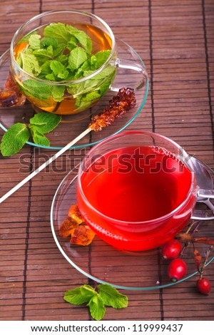 two glasses of fresh herbal rose hip tea and green mint tea with green mint leaf and brown cane sugar on a bamboo board