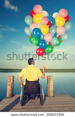 Happy young men in wheelchair holding colorful balloons on wooden boardwalk at lake