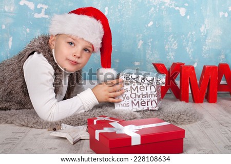 boy in a red Christmas hat lies on fur on the floor and opens his christmas gifts