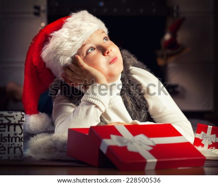 Happy little boy with red santa hat and Christmas present laying by the fireplace
