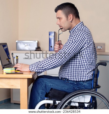 young man in wheelchair in office is using his laptop, disabled young man is surfing in internet