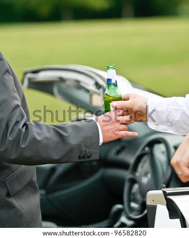 man giving another man a bottle of beer/lager while standing next to a convertible motor vehicle, dangers of drinking and driving