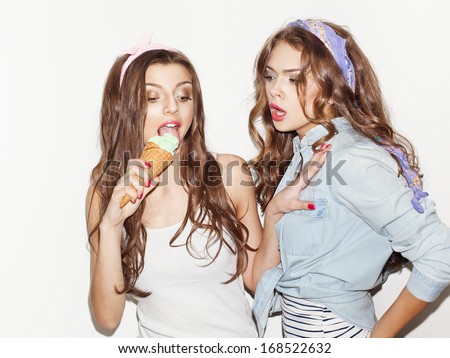 Pretty brunette girls with bright makeup and pink lips having fun. One keeping green ice cream and eating other seems dissatisfied. Inside