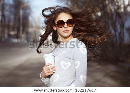 Closeup portrait of beautiful brunette girl with fluttering hair. Keeping takeaway drink in her hand. Walking down the street. City scene. Warm sunny weather. Outdoors