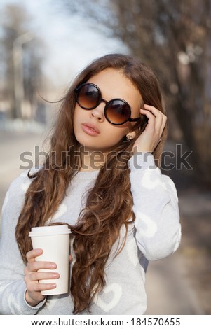 Portrait of a beautiful brunette girl in sunglasses standing on the street. Holding drink in one hand, other next to her face. Warm sunny day. Outdoors