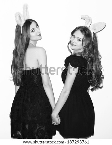 Pretty brunette girl friends standing together and holding hands. Wearing dresses and bunny ears. Looking at camera. Inside