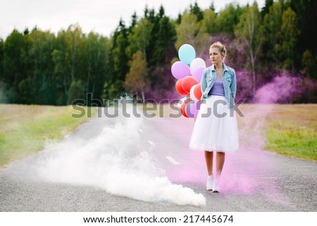 Beautiful blonde girl walking down the road in a countryside, holding color balloons behind her back. Outside