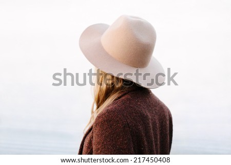 Portrait of blonde girl standing back to camera on lakeside. Wearing hat and jacket. Outside
