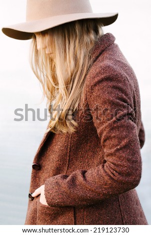 Portrait of a beautiful blonde girl walking down lakeside. Profile to camera. Wearing hat and jacket. Outside