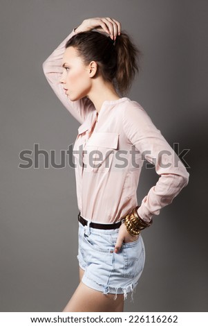 Studio portrait in profile of beautiful young model wearing pink blouse, denim shorts and bracelets. Grey background. Inside