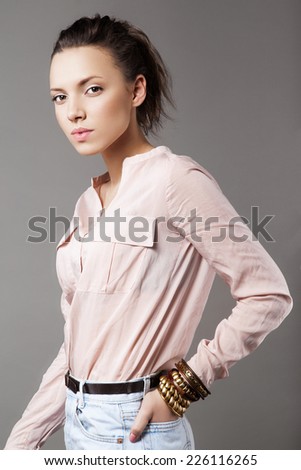 Close up studio portrait of beautiful young model wearing pink blouse, denim shorts and bracelets. Grey background. Inside