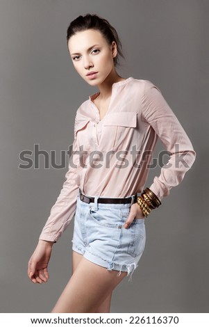 Studio portrait of beautiful young model wearing pink blouse and denim shorts. Grey background. Inside