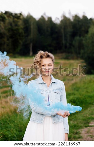 Portrait of beautiful blonde girl keeping flare with blue smoke in her hand. Outdoors