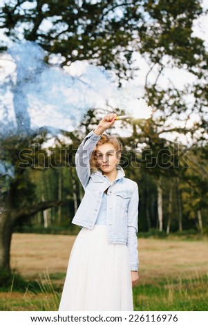 Portrait of beautiful blonde girl keeping flare with blue smoke above her head. Nature background. Outdoors