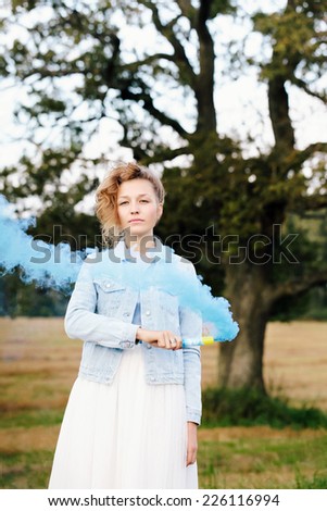 Portrait of beautiful blonde girl keeping blue smoke popper in her hand. Nature background. Outdoors