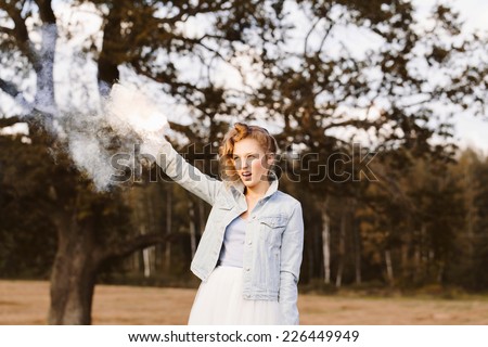 Portrait of beautiful blonde girl wearing casual clothes keeping white smoke flare in her hand. Nature background. Outdoors