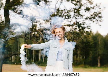 Portrait of beautiful blonde girl wearing casual clothes keeping blue smoke flare in her head. Nature background. Outdoors