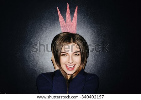 Close up portrait of beautiful smiling girl with perfect skin and nude make up. Pink painted crown on wall. Dark chalk board background.