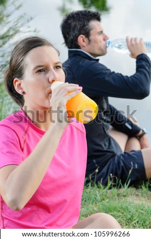 Young athletes drinking and resting after training. Athletes couple sitting in park holding bottle.