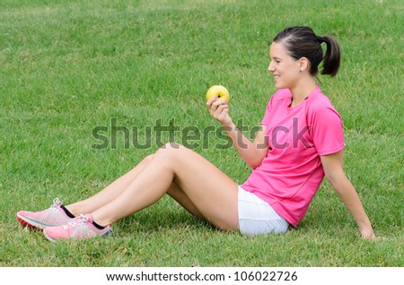 Young sportswoman eating and apple sitting on green grass background. Healthy female athlete lifestyle. Beautiful sporty girl.