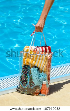 Hand holding handbag at poolside in hot summer day. The bag contains sunglasses, towel, bikini and suntan.