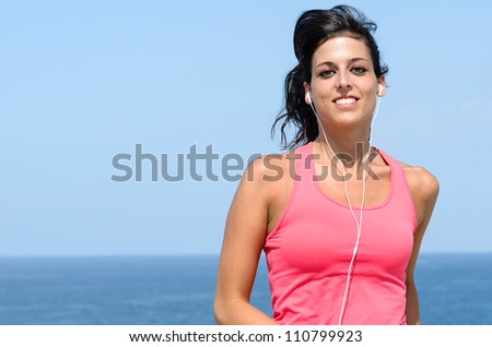 Beautiful sportswoman running outdoors on summer hot day near the sea. Pretty young woman exercising wearing earphones on blue water and sky background.