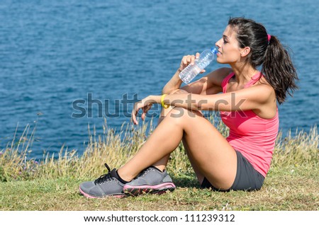 Sport woman resting and drinking water while resting in front of the sea during a hot summer day. She looks to the ocean.