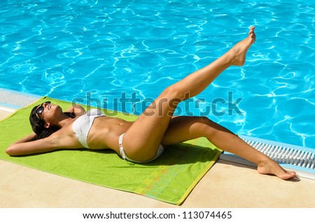 Playful sexy tanned young brunette girl in bikini and sunglasses at poolside sunbathing and relaxing. Caucasian attractive brunette model near the blue water.