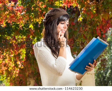 Female teen student thinking with finger in lips and reading outdoors on autumn background