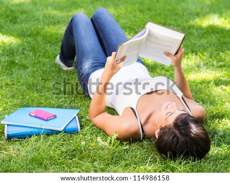 Young brunette female student reading book lying down on grass outside.