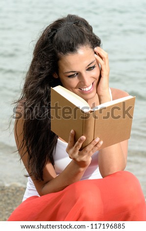 Brunette young woman reading a book and smiling happy on sea background in summer afternoon. She is touching her face with her hand with dreamy looking.